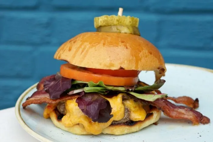 A cheeseburger topped with crispy bacon, leafy greens, tomato slices, and melted cheese on a brioche bun, garnished with sliced pickles and a toothpick, served on a white plate against a blue background.