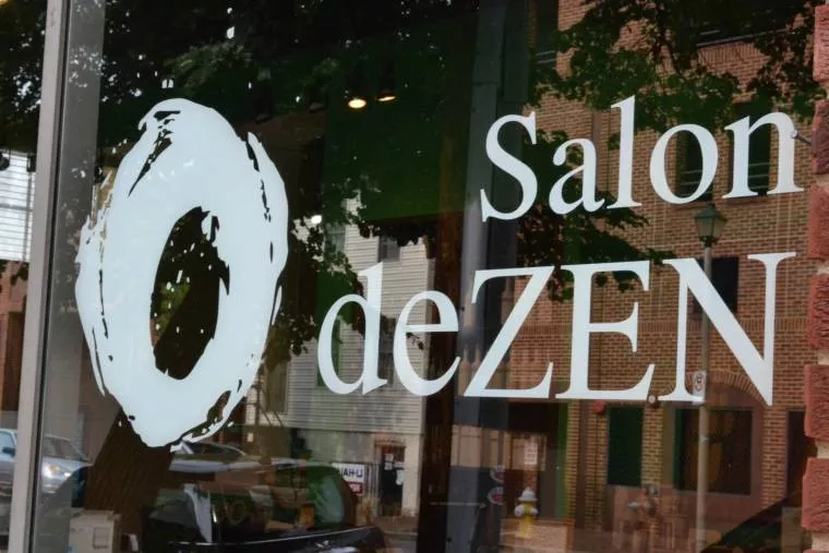 The image shows a glass storefront window with the logo and text Salon deZEN in white, along with a large circular brushstroke design. Reflections of trees, buildings, and cars are visible in the glass.