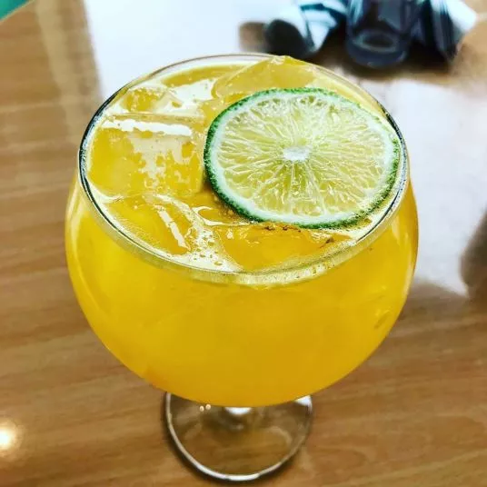 A round glass filled with a bright yellow iced drink, garnished with a slice of lime, sits on a wooden table. A striped napkin and an empty glass are blurred in the background.