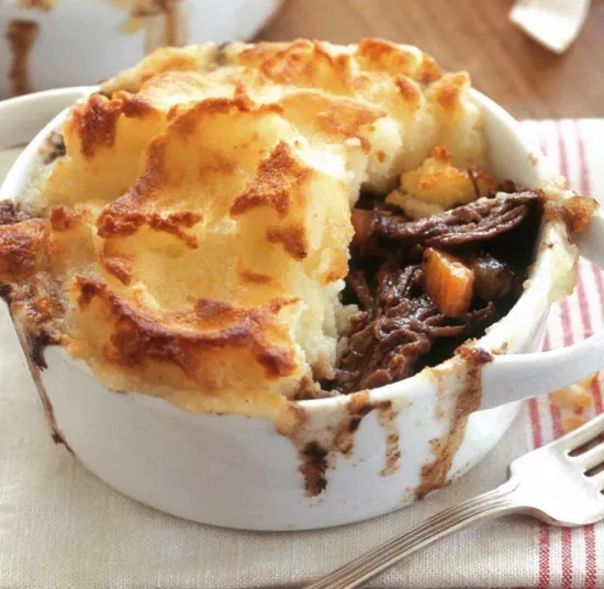 A white ramekin filled with shepherd’s pie featuring a golden-brown mashed potato topping over a filling of shredded meat, carrots, and gravy, with some filling spilling over the edge. A fork and napkin are beside it.