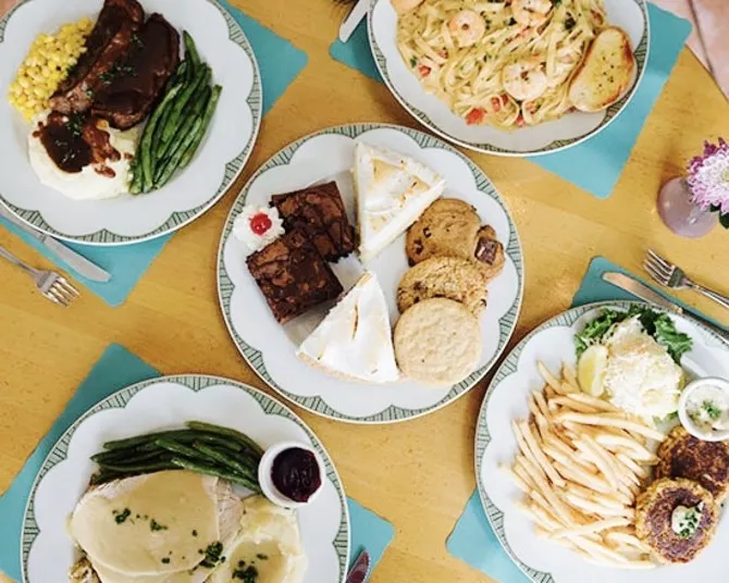 Four plates of food on a wooden table: one with steak, mashed potatoes, corn, and green beans; one with shrimp pasta; one with various desserts; and one with a burger, fries, and a salad.