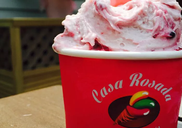 A close-up of a red Casa Rosada cup filled with pink and white swirled gelato, placed on a wooden surface with a blurred background.