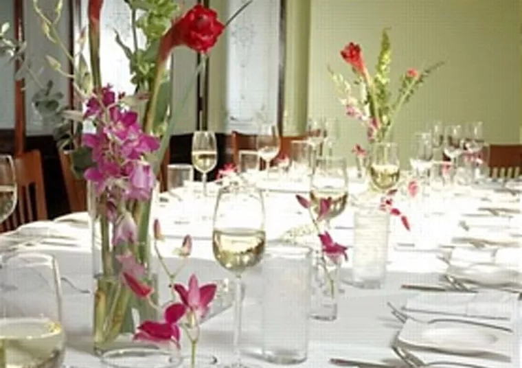 A formal dining table is set with wine glasses, water glasses, white plates, and silverware. Tall vases with red and purple flowers serve as centerpieces along the length of the table.