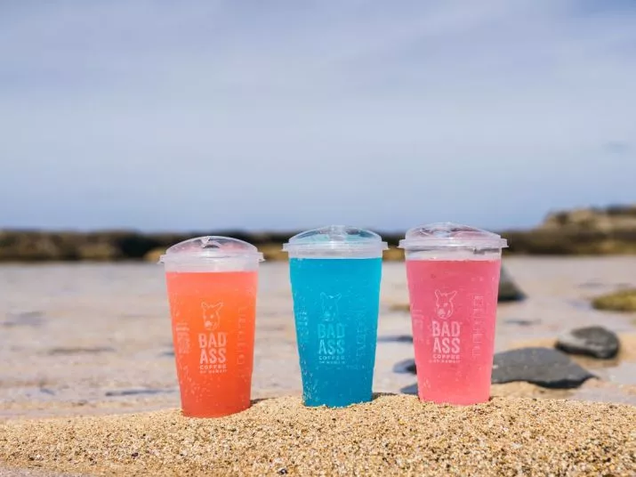 Three colorful drinks in clear cups labeled BAD ASS sit on a sandy beach with rocks and the ocean in the background. The drinks are pink, blue, and pink. The sky is clear and sunny.