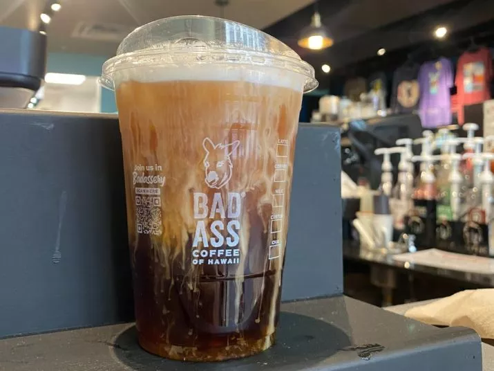 A clear plastic cup of iced coffee with cream swirling through it, branded Bad Ass Coffee of Hawaii, sits on a counter in a coffee shop with syrup bottles and merchandise visible in the background.