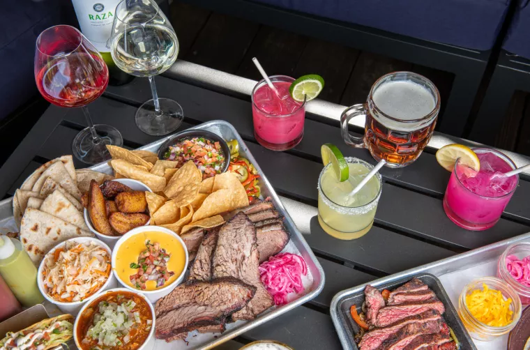A table set with trays of smoked meats, tortillas, chips, queso, slaw, and plantains, surrounded by colorful cocktails, wine, and beer, creating a vibrant, inviting meal outdoors.