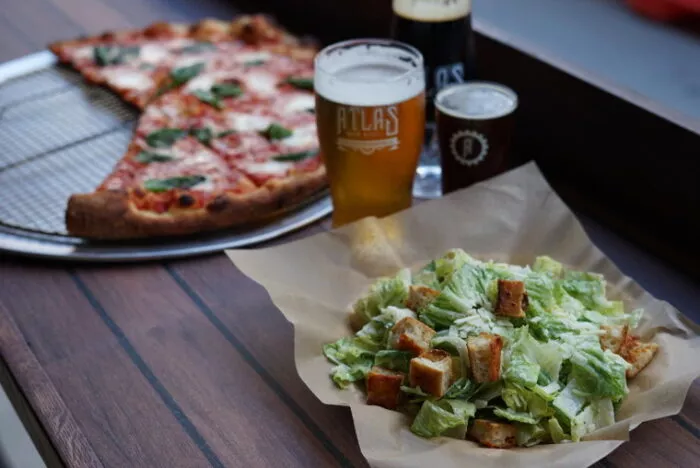 A plate of Caesar salad with croutons sits on a wooden table next to two large slices of pizza on a tray and two glasses of beer.