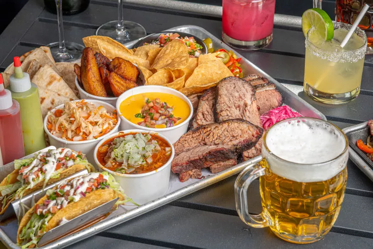 A tray filled with assorted foods, including sliced brisket, tacos, fried plantains, chips with cheese dip, coleslaw, and bowls of salsa, surrounded by various drinks like beer and cocktails.