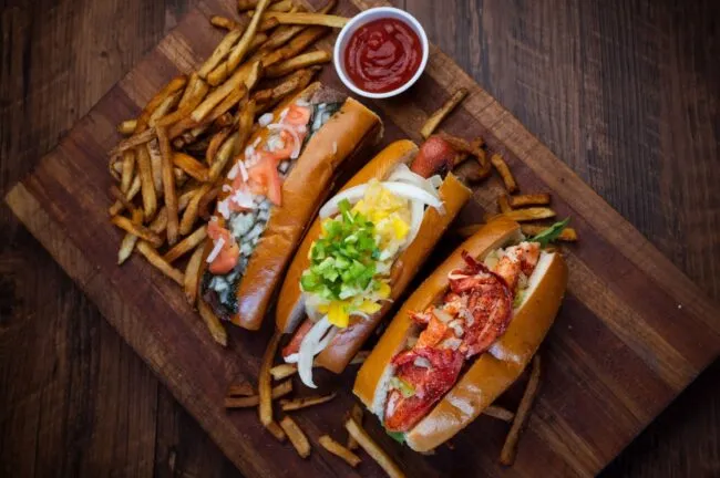 Three gourmet hot dogs with various toppings and a pile of French fries are arranged on a wooden board, with a small cup of ketchup for dipping in the upper center of the image.