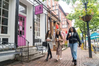 Three women walk along a brick sidewalk, each holding a drink, passing shops including one with a pink door and a sign that reads 3 Sisters. The atmosphere is casual and friendly on a tree-lined street.