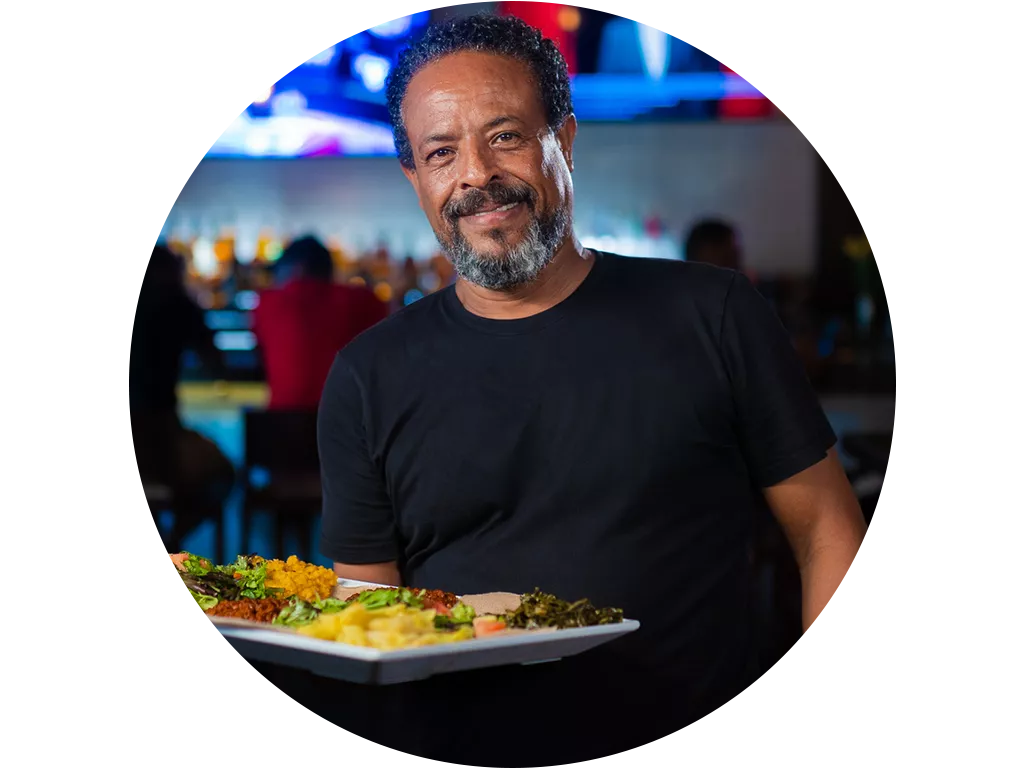 A smiling man in a black t-shirt holds a large plate of assorted food in a lively restaurant or bar with people and colorful lights in the background.