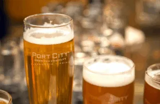 Two glasses of beer, one taller and one shorter, both with foamy heads, display the Port City Brewing Company Alexandria, Virginia logo. Blurred glasses and a warm background are visible behind them.