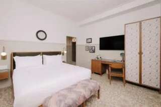 A modern hotel room with a king-size bed, patterned bench, desk with chair, large flat-screen TV, wardrobe, wall art, and neutral-colored decor. The room has soft lighting and a contemporary, cozy feel.