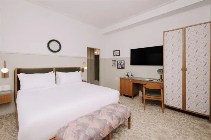 A modern hotel room with a king-size bed, patterned bench, desk with chair, large flat-screen TV, wardrobe, wall art, and neutral-colored decor. The room has soft lighting and a contemporary, cozy feel.