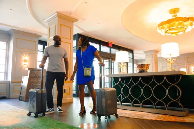 A man and woman, each with a rolling suitcase, smile at each other while walking through a stylish hotel lobby with elegant decor and warm lighting.