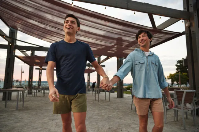 Two young men are smiling and holding hands while walking outside under a canopy structure at sunset. They appear happy and relaxed, dressed casually in shorts and shirts. Empty chairs and tables are visible in the background.