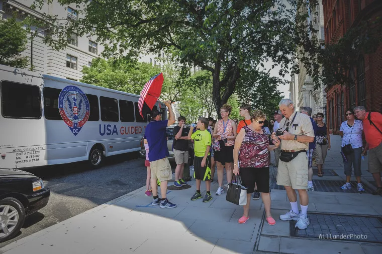 A tour guide holding a red umbrella leads a group of tourists on a sunny sidewalk near a USA Guided tour bus. The group listens attentively and stands near trees and city buildings.