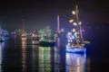 Boats decorated with colorful holiday lights and ornaments float on calm water at night, reflecting vibrant colors on the surface during a festive boat parade.