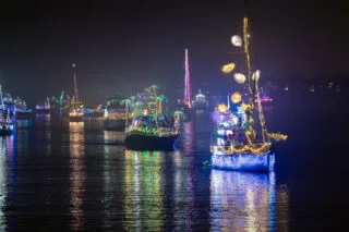Boats decorated with colorful holiday lights and ornaments float on calm water at night, reflecting vibrant colors on the surface during a festive boat parade.
