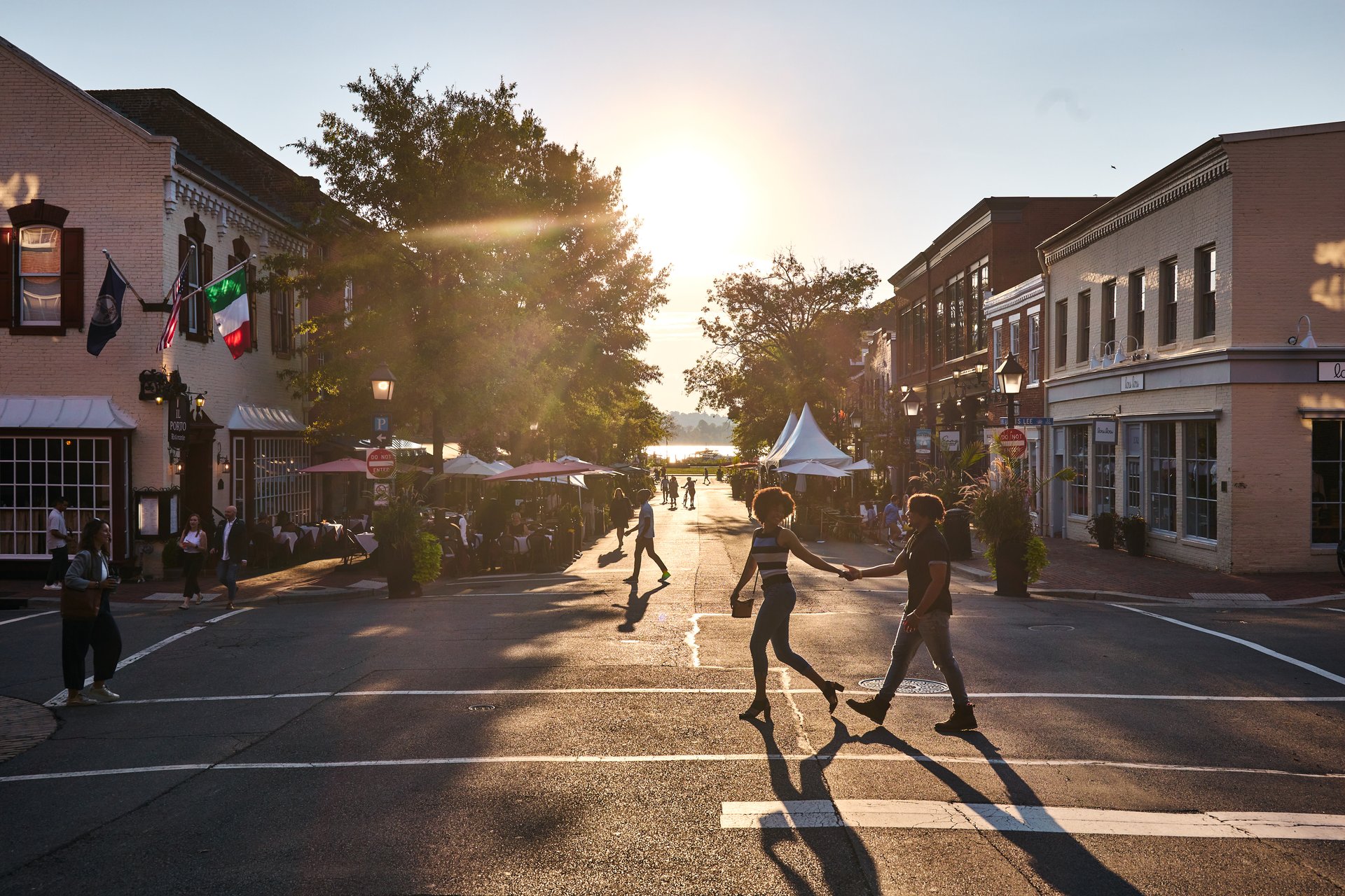 Two people holding hands cross a street in a lively town at sunset, with outdoor diners, people walking, and sunlight streaming between historic buildings. Trees and tents line the background.