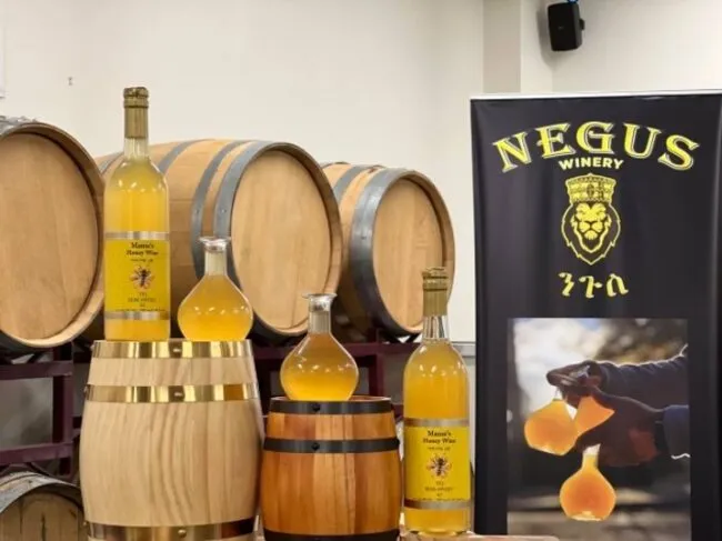 Bottles and carafes of honey wine are displayed on wooden barrels next to a Negus Winery banner featuring a lion logo and Amharic text. A photo on the banner shows honey wine being poured into glasses.