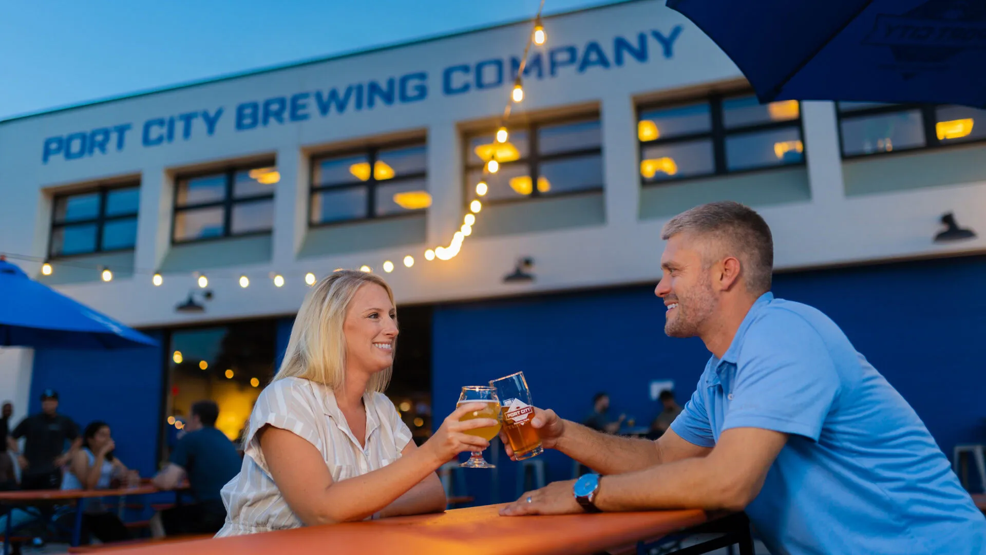 A woman and man sit at an outdoor table, smiling and clinking beer glasses at Port City Brewing Company, with string lights and people in the background during the evening.