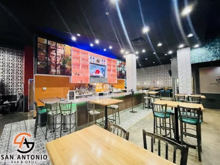 Brightly lit modern bar and grill with wooden tables, green chairs, a colorful mural above the bar, decorative tile walls, and the San Antonio Bar & Grill logo in the bottom left corner.