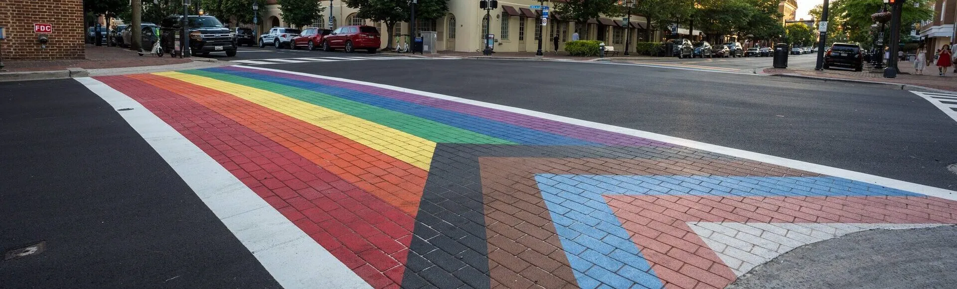 A crosswalk painted with the Progress Pride flag colors spans a street in an urban area with brick buildings, trees, parked cars, and traffic lights visible in the background.