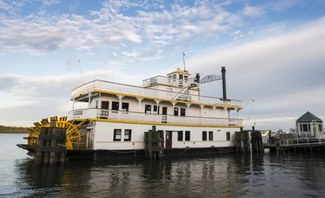 A white and yellow paddlewheel riverboat is docked at a pier on calm water under a partly cloudy sky.