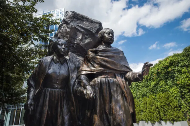A bronze statue of two women standing side by side, holding hands, with a large rock behind them. Trees and a building covered with ivy are visible in the background under a partly cloudy sky.