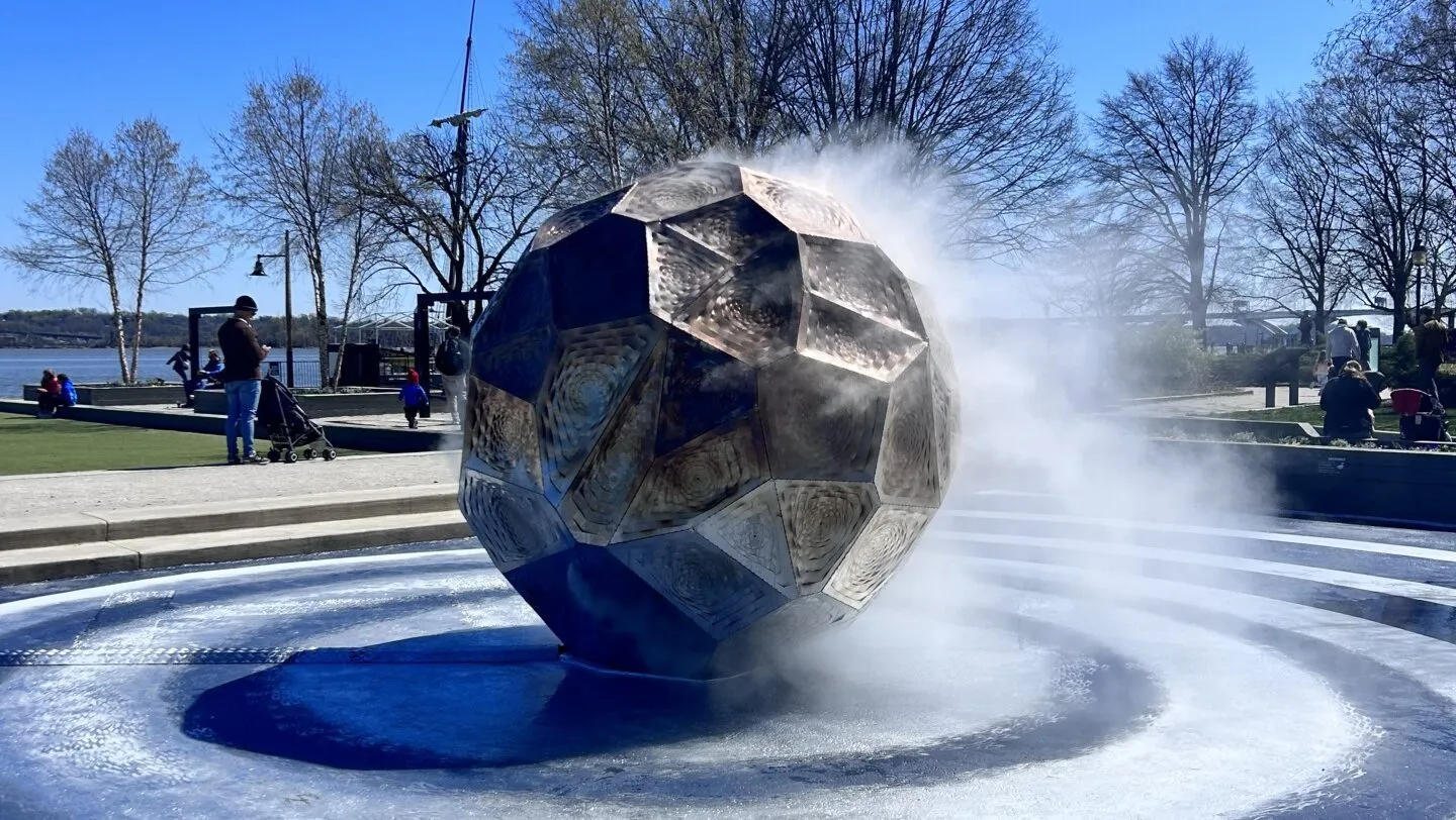 A large metallic soccer ball sculpture emits mist in a circular fountain, with people walking and trees in the background under a clear blue sky.