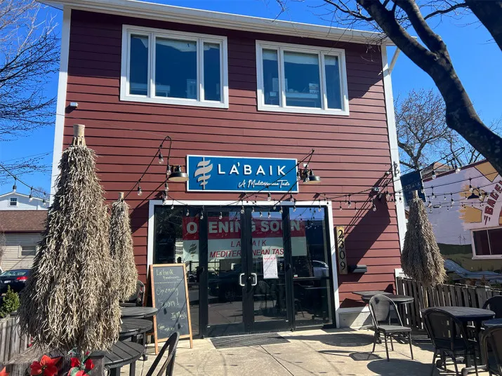A red two-story restaurant with large windows, a blue sign reading “Labaik: A Mediterranean Tab” above the entrance, outdoor tables, and two straw umbrellas on a sunny day.