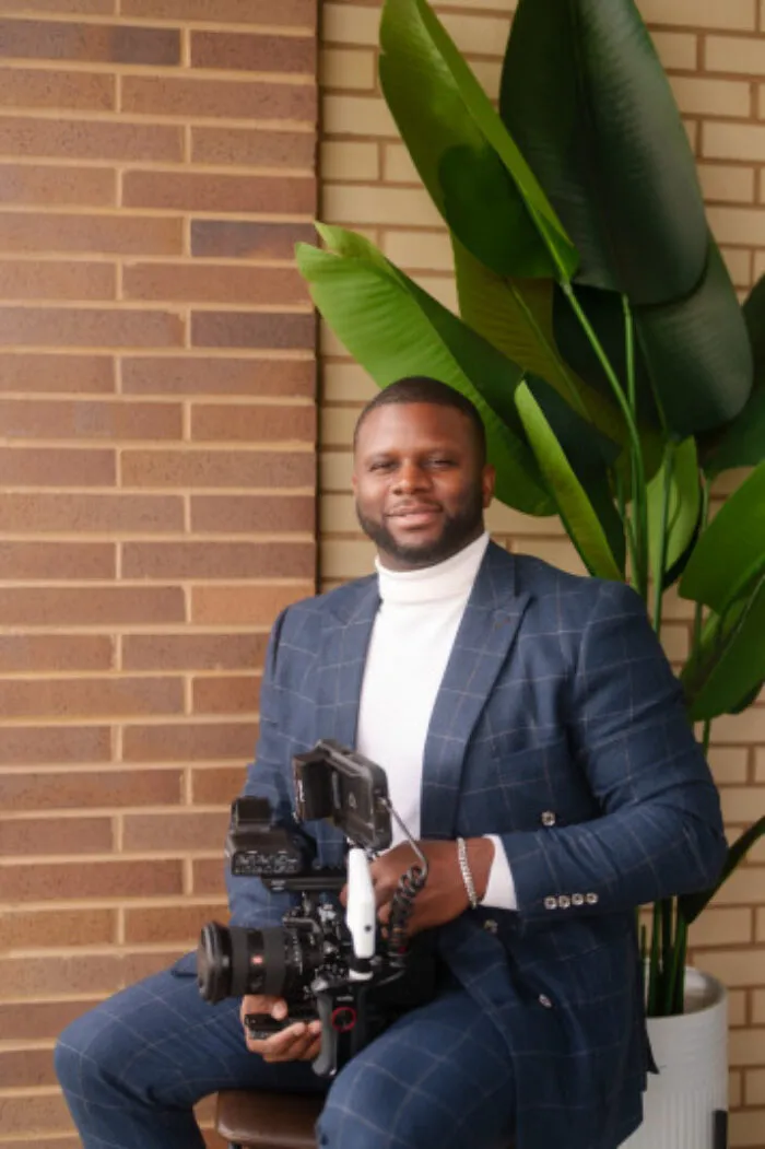 A person in a blue checked suit and white turtleneck sits on a stool, smiling and holding a professional video camera, with a large green plant and brick wall in the background.