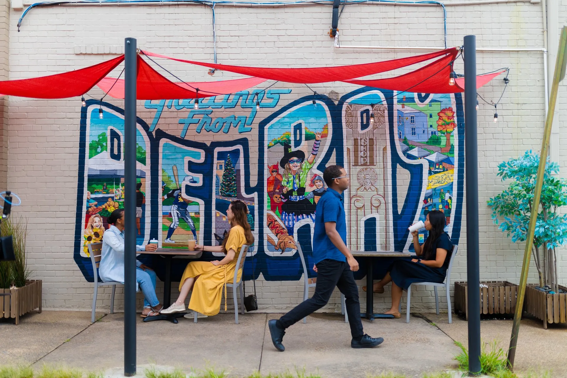 Three people sit at outdoor tables under red canopies, talking and drinking, while a person walks by. The background features a colorful Greetings from Bel Air mural on a brick wall.