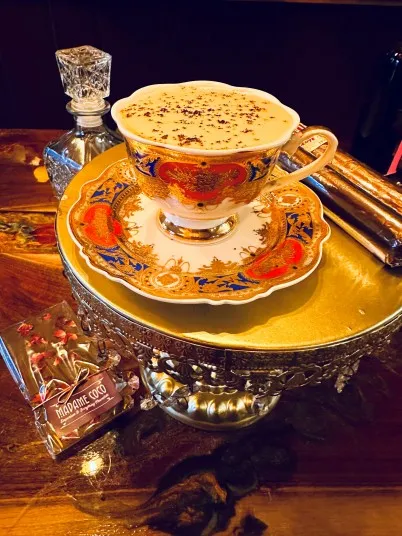 A decorative teacup filled with a frothy drink sits on an ornate saucer atop a gold stand. Nearby are a glass decanter, two cinnamon sticks, and a small bag of Madame Coco chocolates.