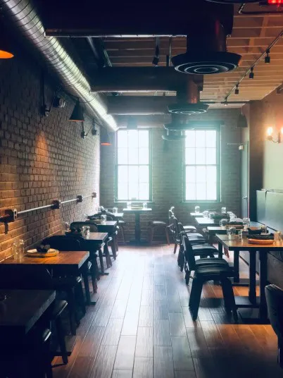 A cozy restaurant dining area with exposed brick walls, wooden tables and chairs, place settings, and large windows letting in natural light. The space has a warm, inviting atmosphere with industrial decor elements.