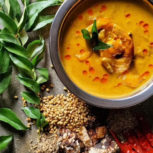 A bowl of yellow curry with shrimp, garnished with curry leaves and chili oil, sits beside fresh curry leaves, dried red chilies, coriander seeds, cumin seeds, and tamarind pieces.