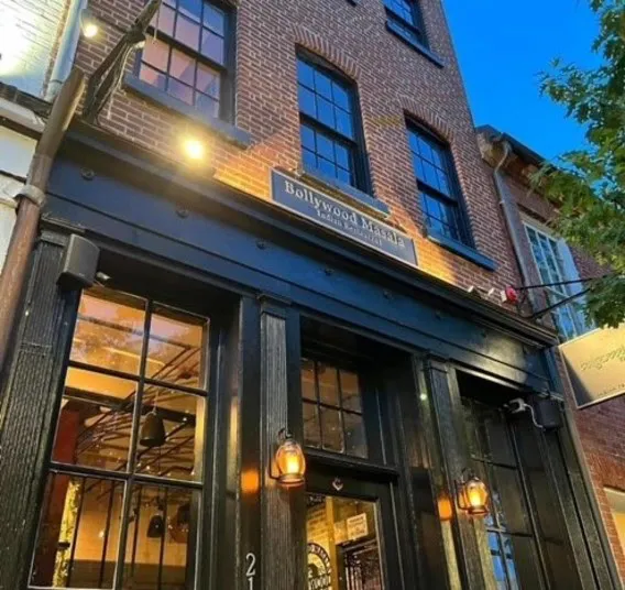 A brick building with large black-framed windows and a sign reading Bollywood Masala above a dark entryway, warmly lit by exterior lanterns, taken during dusk with a deep blue sky overhead.