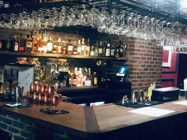 A cozy bar with a wooden counter, shelves stocked with various bottles, hanging glassware overhead, and a brick wall background. Several copper mugs and bar tools are arranged on the counter.