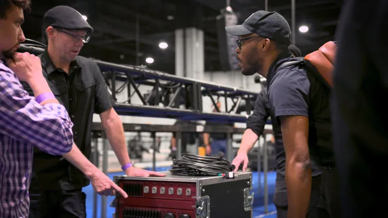 Two men in black clothing and hats discuss while standing next to audio or lighting equipment at an indoor event setup. One points at a control panel, cables are visible on the equipment, and another person listens nearby.