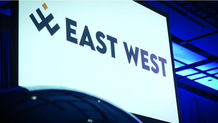 A large screen displays the text EAST WEST with a stylized logo on the left, set in a conference room with blue lighting.