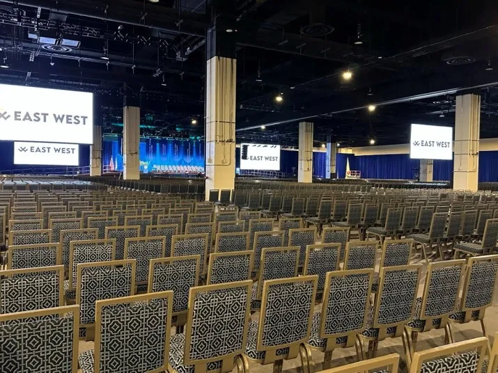 Rows of patterned chairs are arranged in a large conference hall with several large screens displaying the words EAST WEST. The stage is visible in the distance, framed by blue curtains. The hall is empty.