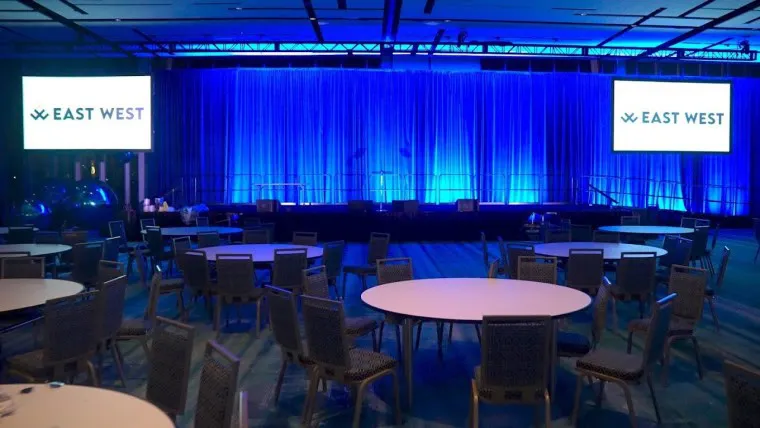 A large event space with round tables and chairs facing a stage with blue lighting. Two screens display the EAST WEST logo on either side of the stage. The room appears empty and ready for attendees.