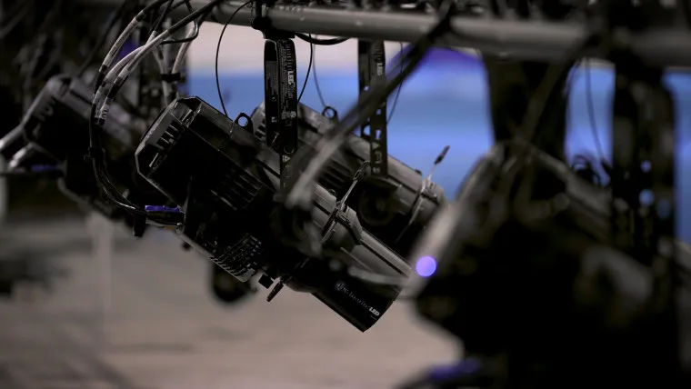 Close-up of several stage lights mounted on a metal rig, with cables hanging around them. The background is blurred, featuring cool blue and gray tones.