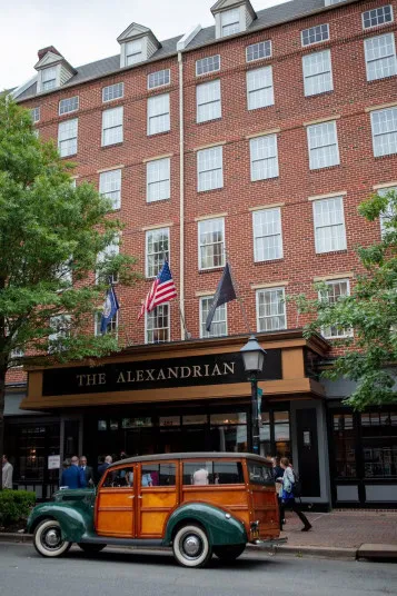 A vintage wood-paneled car is parked in front of The Alexandrian hotel, a red-brick building with large windows and three flags above the entrance. Trees and a few pedestrians are visible nearby.