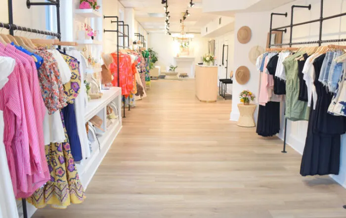 A bright, modern boutique with light wood floors displays colorful dresses and casual clothing on racks along both walls. Hats and accessories are arranged neatly; flowers add decorative touches.