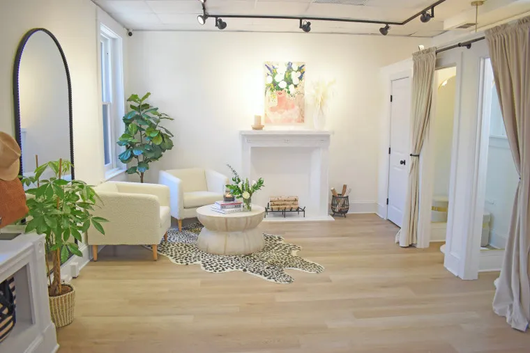 Bright, modern living room with light wood floors, white walls, a round coffee table, cozy armchairs, green plants, a decorative rug, and a fireplace with artwork above it. Curtains enclose a small side area.