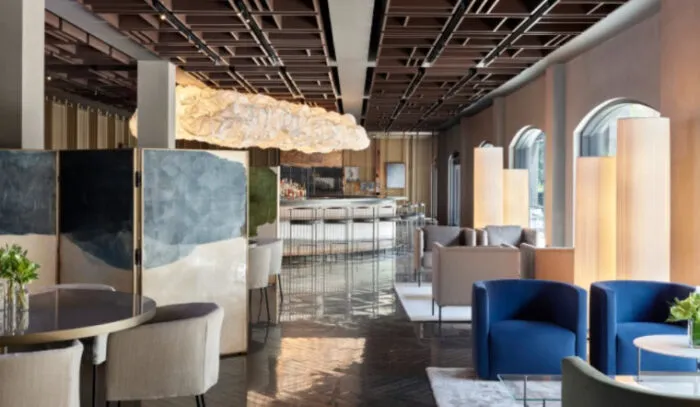 A modern lounge with stylish seating, a sculptural ceiling light, and a sleek bar in the background. Large windows let in natural light, and decorative screens and plants add to the contemporary decor.