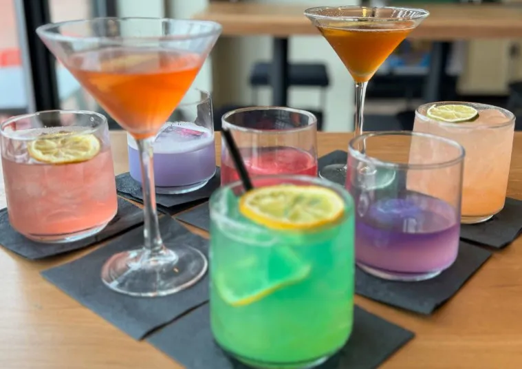 A variety of colorful cocktails, including martinis and drinks garnished with lime slices, are arranged on black napkins atop a wooden table in a modern indoor setting.