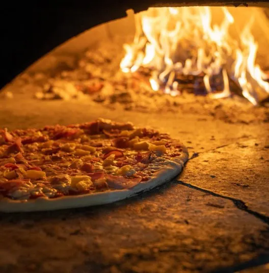 A pizza topped with cheese and pineapple bakes inside a stone wood-fired oven, with flames burning in the background.